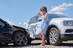 A woman in a blue dress stands by her white SUV and a black car that T-boned it. She talks on her cell phone and holds her head, wondering what to do after a car accident in Las Vegas.