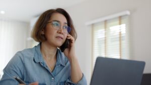 A woman sits at her computer, phone to her ear, looking worried, and wonders how to report elder abuse in California.