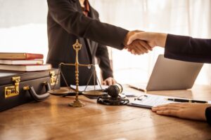 Lawyer shaking hands with a client over a desk with scales of justice, a gavel, a briefcase, and a document